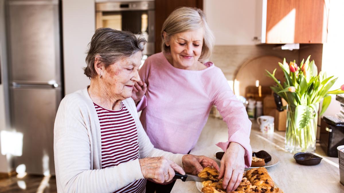 Zwei Personen in einer Küche. Die ältere Frau im Vordergrund schneidet Kuchen.